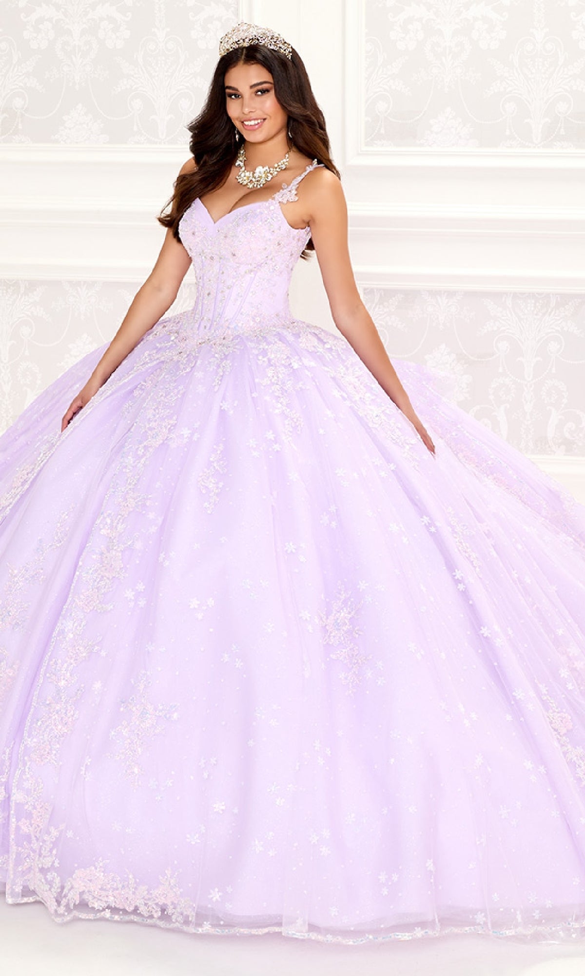 A woman in the Quinceanera Dress PR30087 By Princesa, featuring light purple floral embellishments and a corset bodice, with a tiara and statement necklace, stands in an ornate white room.